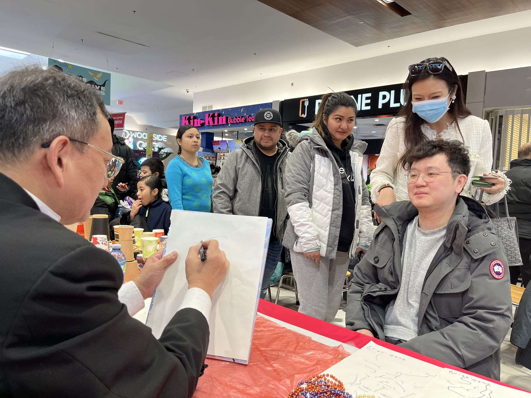 A person signing a book with a group of people in a mall

Description automatically generated